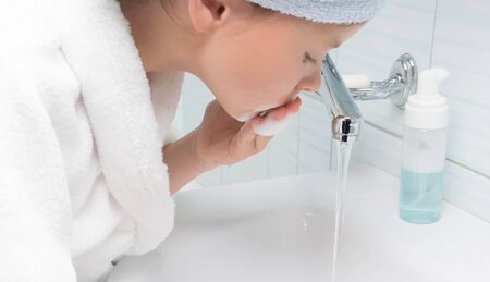 in the bathroom, a girl in a white bathrobe, lathers her face with foam for cleaning, against the...