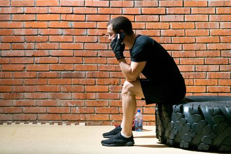 a man, sitting on a black wheel, in between sports and talking on the phone, against a brick wallの写真素材