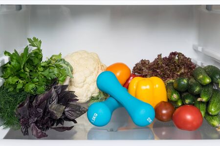 slimming concept: dumbbells and fresh vegetables on a shelf in the refrigeratorの写真素材