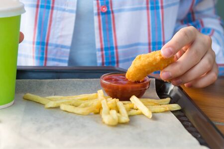 children's hand holds chicken nuggets and dips it in ketchup, next to it lies french fries