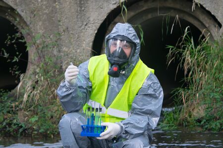 specialist in a gray protective suit and mask, on the pond, takes a sample of liquid in test tubes,...