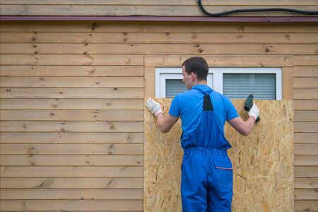 a worker in a blue uniform, blocks the window of the house with a protective shield made of wood,...