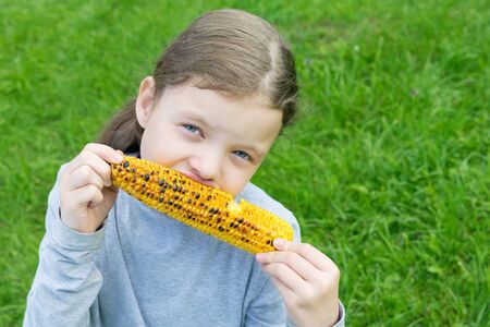 little girl outdoors sitting on the grass and eating grilled corncob