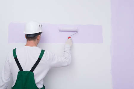 against the background of a painted wall, a master in protective clothing applies paint with a rollerの写真素材
