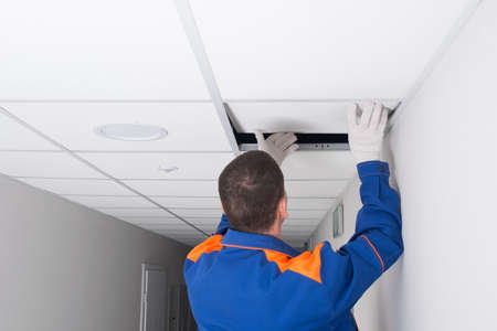 a worker in protective clothing opens a suspended ceiling plate for repairsの写真素材