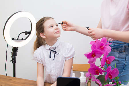 during the break, under the light of the ring lamp, the blogger girl, at the table, is preparing for a video chat, the assistant puts mascara on her eyelashesの写真素材