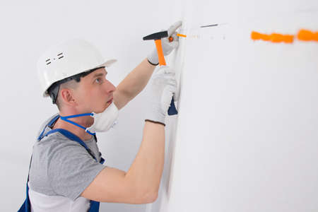 in the white wall, holes are made and orange dowels are hammered with a hammer, a worker in a protective helmetの写真素材