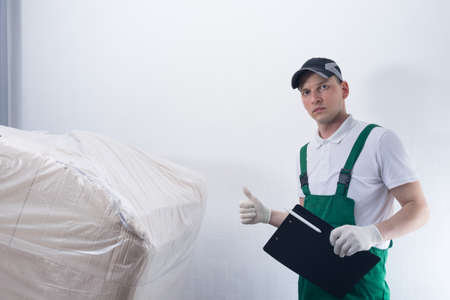 a man in a work uniform delivered a large box to the address, shows a thumbs up, holding documents in his handの写真素材