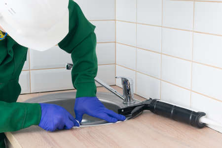 the master in protective gloves, wipes off the excess silicone, after installing the metal sink of the wooden countertopの写真素材