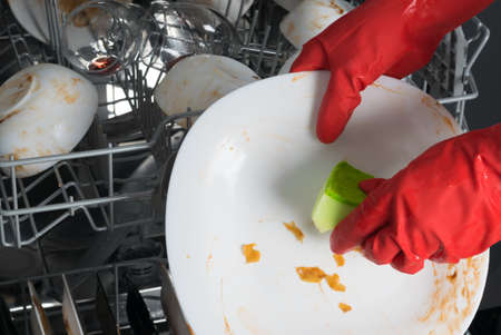 a hand in a red protective glove, cleans white plates of large food debris, for putting in the dishwasher, close-upの写真素材