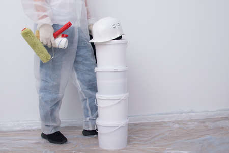 a man in protective clothing, with paint rollers in his hands, put four buckets on top of each other, against the background of a white wall, there is a place for the inscriptionの写真素材