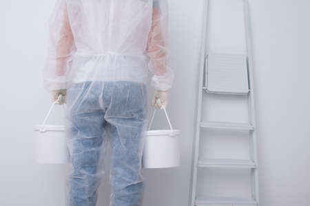 a man in protective clothing, bringing buckets of paint, against the background of a white wall and a stepladder, rear viewの写真素材