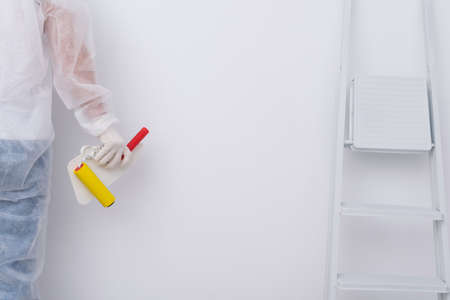 a construction worker in protective clothing holds a spatula and a roller in his hand, against a white wallの写真素材