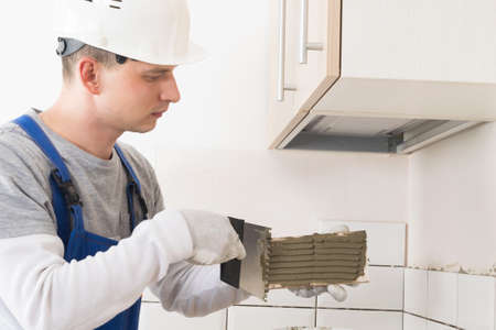 worker in overall applies glue to the tile with a spatula to lay it on the wall in the roomの写真素材
