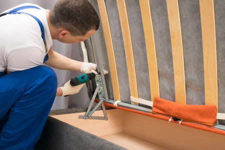 the deliveryman assembles upholstered furniture with the help of an electric screwdriver at the buyer's houseの写真素材