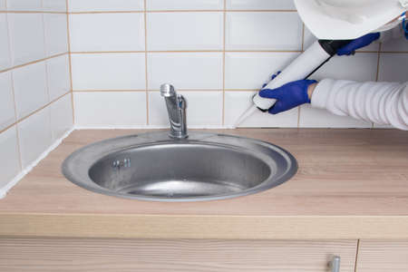 master of interior decoration, smears the joint of the tile and the kitchen countertop with white silicone instead of the skirting board, close-up, against the background of the sinkの写真素材