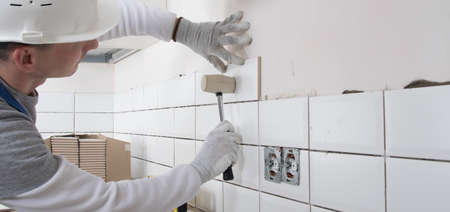 a builder in a protective helmet, using a rubber hammer, corrects the correct installation of tilesの写真素材