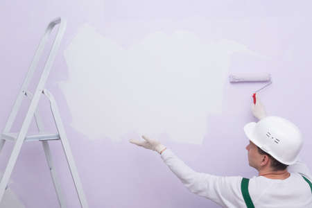 a construction worker in protective clothing and a hard hat, applies paint with a roller and shows a clean place on the wallの写真素材