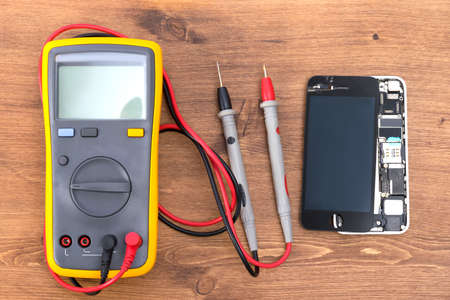 on a wooden table there is an electric tester equipment and a phone with a removed liquid crystal screen for repairの写真素材