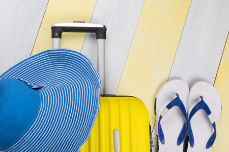 on a light wooden striped background there is a yellow suitcase and a set of things for recreation a hat shoes flip flops, there is a place for the inscriptionの写真素材