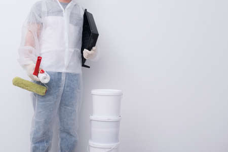 a man in protective clothing, with paint rollers in his hands, put three buckets on top of each other, against the background of a white wall, there is a place for the inscriptionの写真素材