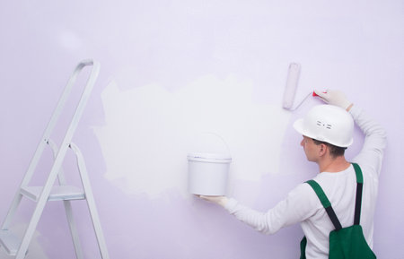 a construction worker in protective clothing and a helmet, applies paint with a roller and holds a bucket in his hand, next to a folding ladderの写真素材