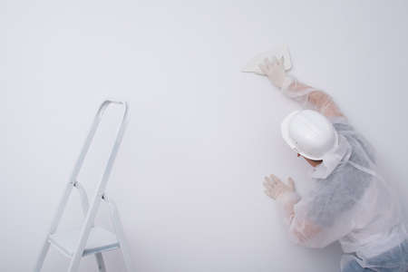 a construction worker in protective clothing and a hard hat, levels the wallpaper with a plastic spatula, to remove excess glue from the walls, rear viewの写真素材