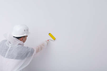 a construction worker in protective clothing and a helmet, smoothes the wallpaper on the white wall with a special roller, there is a place for the inscription, a rear viewの写真素材