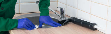 master in protective gloves, wipes off excess silicone, after installing the metal sink of the wooden countertop, close-upの写真素材
