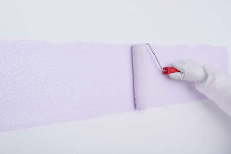 a painter's hand in protective gloves, with a large roller, painting a white wall in a light purple...