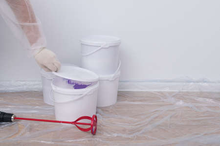 a construction worker's hand in protective clothing opens a bucket of paint to stir it, against a white wallの写真素材