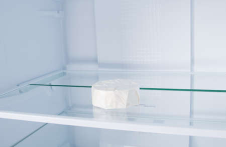 against the background of a white refrigerator, brie cheese in a closed package is lying on a glass shelf, close-upの写真素材