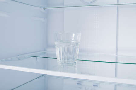 against the background of a white refrigerator, there is a glass of water on a glass shelf, close-upの写真素材