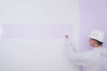 a painter in protective clothing, paints a white wall in a light purple color, there is a place to writeの写真素材