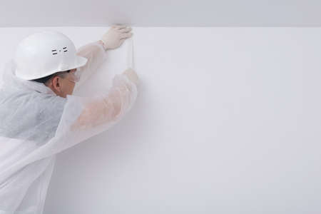 a construction worker in protective clothing and a hard hat, glues paint tape to create a multi-colored interior, on a white wallの写真素材