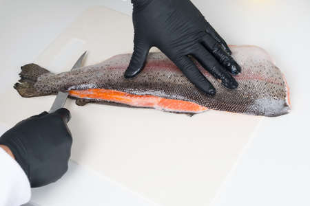 the process of cutting red trout, by a chef in black gloves, with a sharp knife on a white table, close-upの写真素材