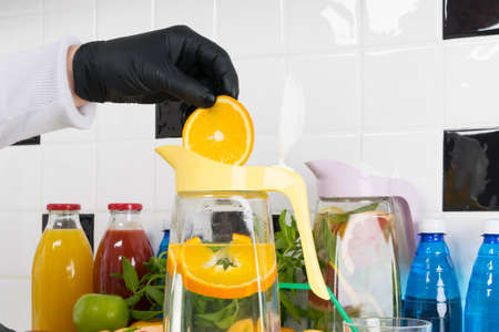 the cook, wearing protective gloves, puts chopped citrus fruits and mint in a glass jug to prepare a refreshing drinkの写真素材
