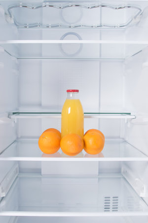 against the background of a white refrigerator, there is a bottle of juice and oranges on a glass shelfの写真素材