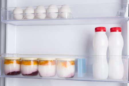 in the door of an open white refrigerator, on the shelf are yoghurts and eggsの写真素材