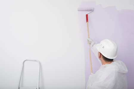 a worker in a hard hat paints a white wall with a roller in lilac colorの写真素材