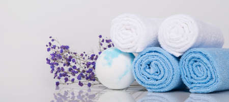 on a white table with a reflection, towels, four rolled-up spa treatments and blue bath salts in the form of a ball, next to flowers for decoration, close-upの写真素材