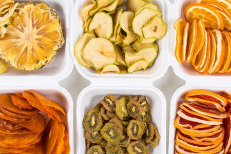 in plastic white containers there are dried fruits in slices, pineapple, persimmon, kiwi, apple, orange and grapefruit, close-upの写真素材