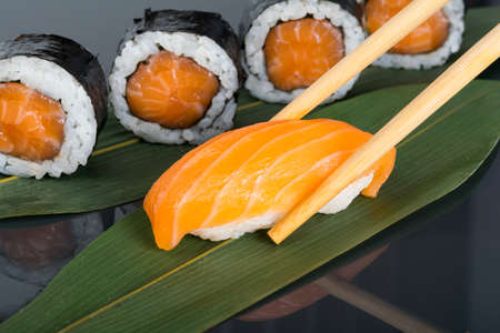 close-up, on a dark table with a reflection, on a green bamboo leaf, wooden sticks holding sushi, surrounded by Japanese salmon rollsの写真素材
