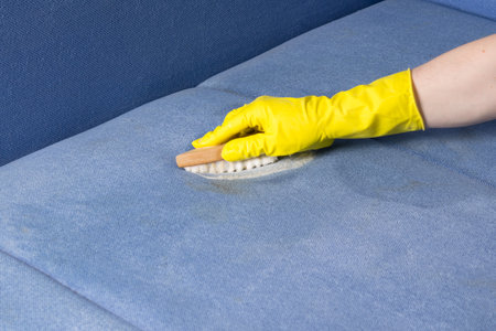 close-up of a hand in yellow gloves cleaning the blue sofa cloth using a brush and foamの写真素材