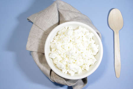 Cottage cheese in white bowl on blue table with spoon and napkin. Top view. Horizontal orientation.