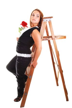 Young, lovely girl in black pants and top, with white shirt holding a red rose in her hand and leaning on the stepladder, for light gray background.の写真素材