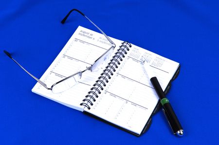 A day timer and personal planer with glasses and a pen on royal blue background.の写真素材