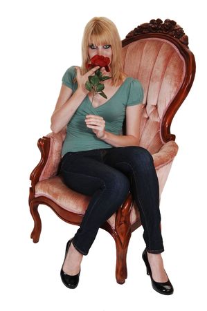 A young pretty woman in jeans and green sweater sitting in an old fashion pink armchair with a rose in her hand, for white background.の写真素材