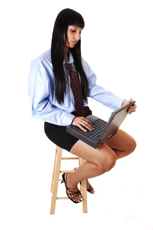 A nice young woman sitting on a chair with a laptop on her lap, doing some work, in shorts and a blue shirt with tie and long black hair, over white.の写真素材
