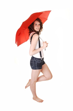 A young pretty woman in jeans shorts with suspender and a white t-shirt standing bare feet and holding an umbrella over her shoulder, over white.の写真素材